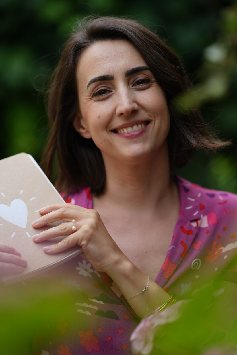 Femme souriante tenant un carnet, entourée de feuillages