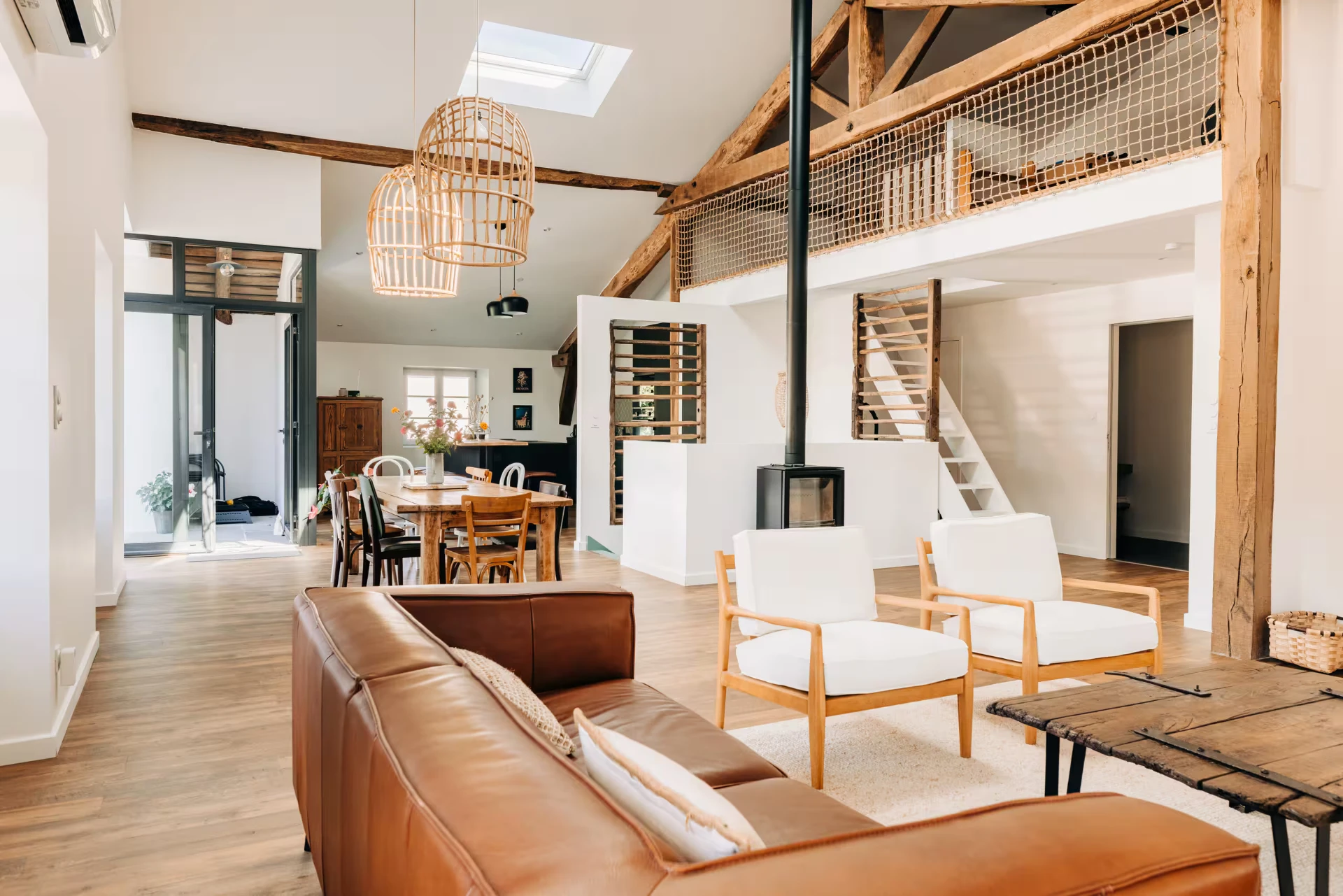 Salon lumineux de l'ancien fenil : charpente apparente, poêle à bois, canapé en cuir et grande table