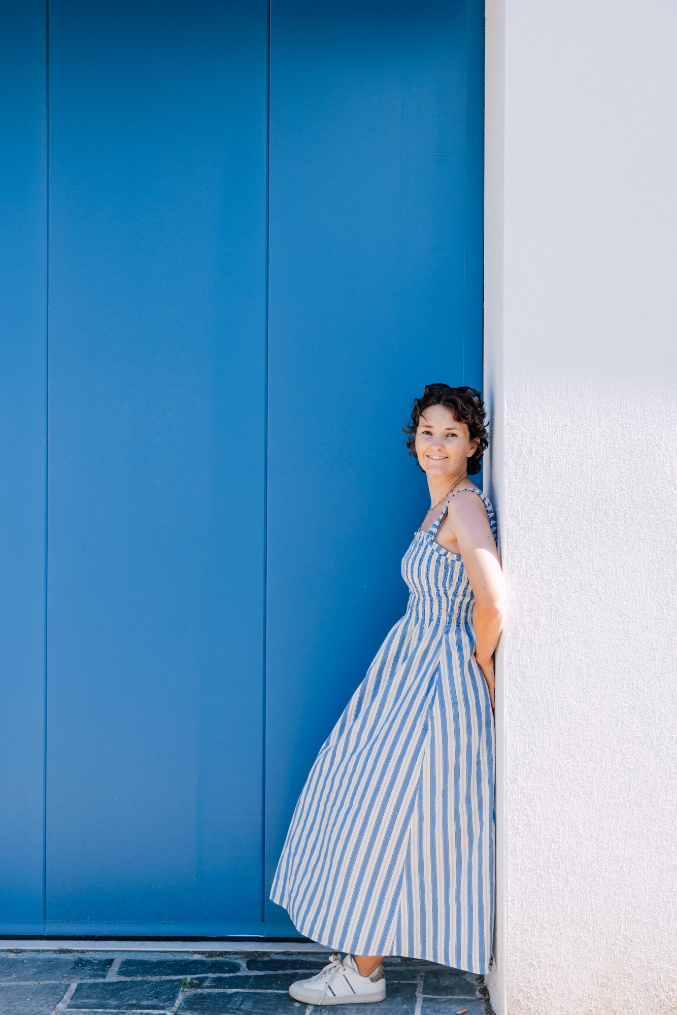 Femme en robe rayée adossée à une porte bleue en plein soleil